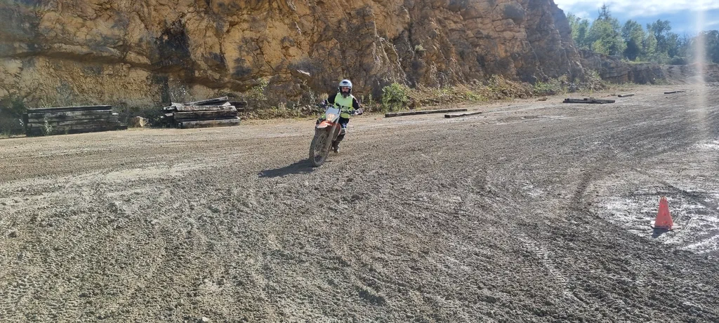 Un pilote en gilet jaune sur une moto d'enduro au fond d'une carrière, au pied de la falaise ; au premier plan, un sol de gravier humide strié de traces de pneus et un cône orange.